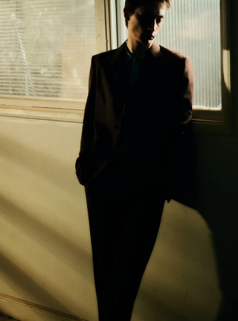 Fashionable adult male in a dark suit stands by a window with sunlight. Dramatic shadows and the model's pose create a moody atmosphere.