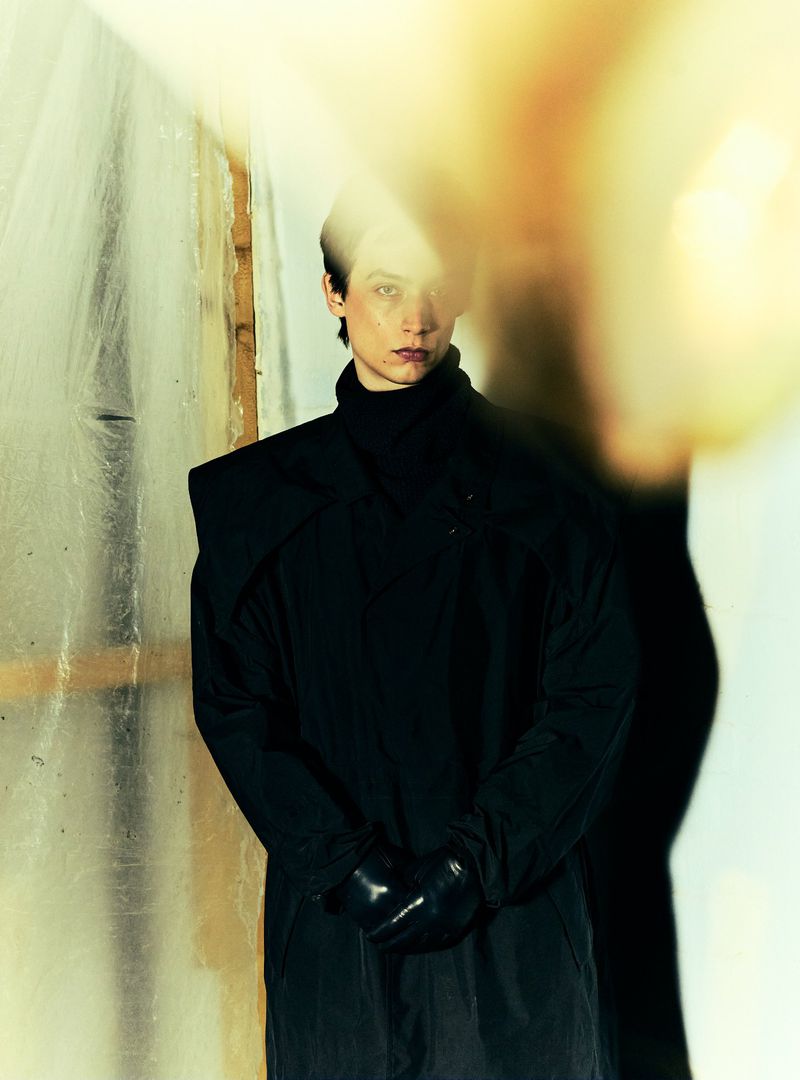 Adult female with dark hair in black coat and gloves, stands indoors against textured wall, backlit by blurred yellow light.