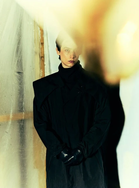 Adult female with dark hair in black coat and gloves, stands indoors against textured wall, backlit by blurred yellow light.
