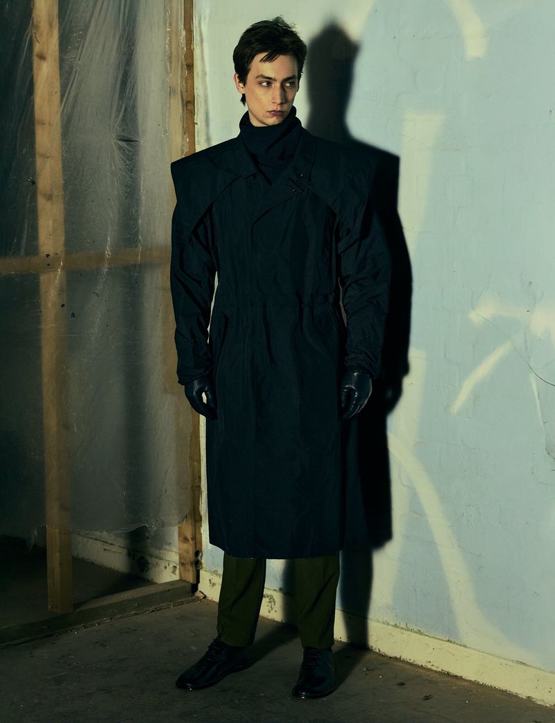 Fashionable young adult male posing indoors. Man wears a long black coat with green pants and black shoes in a dimly lit corner.
