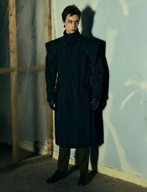 Fashionable young adult male posing indoors. Man wears a long black coat with green pants and black shoes in a dimly lit corner.
