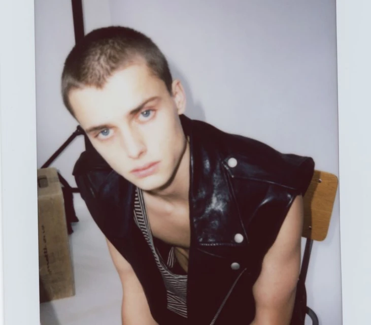 Young adult man with short hair and blue eyes wearing a black leather vest poses indoors against a plain white wall in a fashion style.