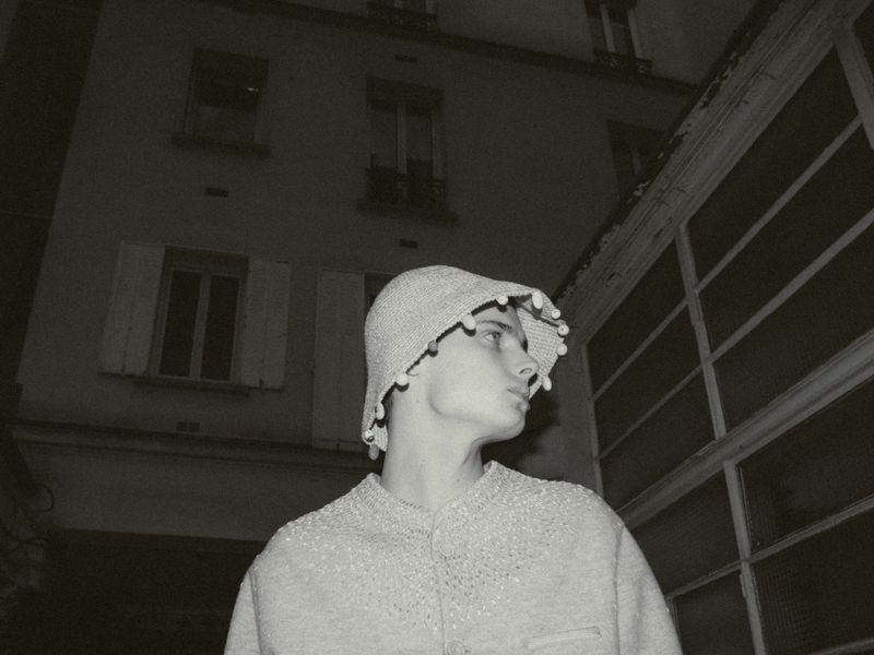 Monochrome portrait of a young person in a decorative pom-pom hat and sweater, set against an urban building exterior at dusk.