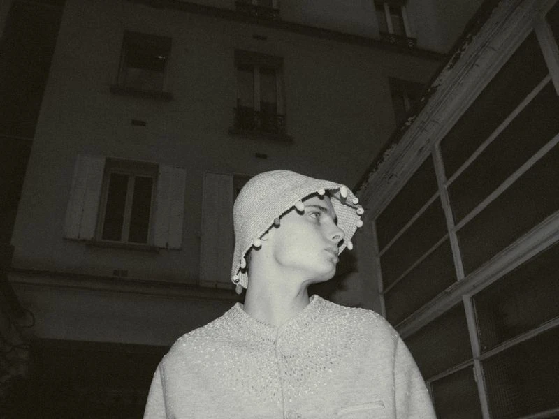Monochrome portrait of a young person in a decorative pom-pom hat and sweater, set against an urban building exterior at dusk.