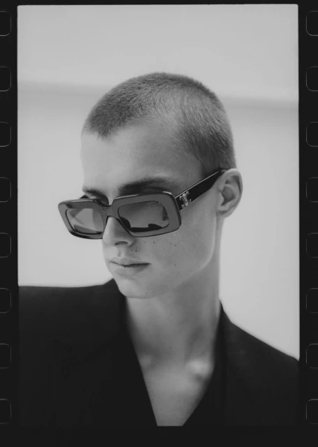 Monochrome portrait of a person with a buzzcut wearing chunky sunglasses and a dark blazer against a plain, light background.