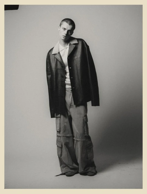 A young adult man poses in an oversized jacket and cargo pants in a studio, creating a high-fashion editorial shot. 