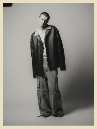 A young adult man poses in an oversized jacket and cargo pants in a studio, creating a high-fashion editorial shot. 