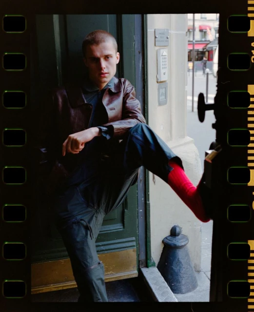 A young adult poses with one leg up in front of a dark green door and city street in the background. He is wearing a brown jacket and dark pants.