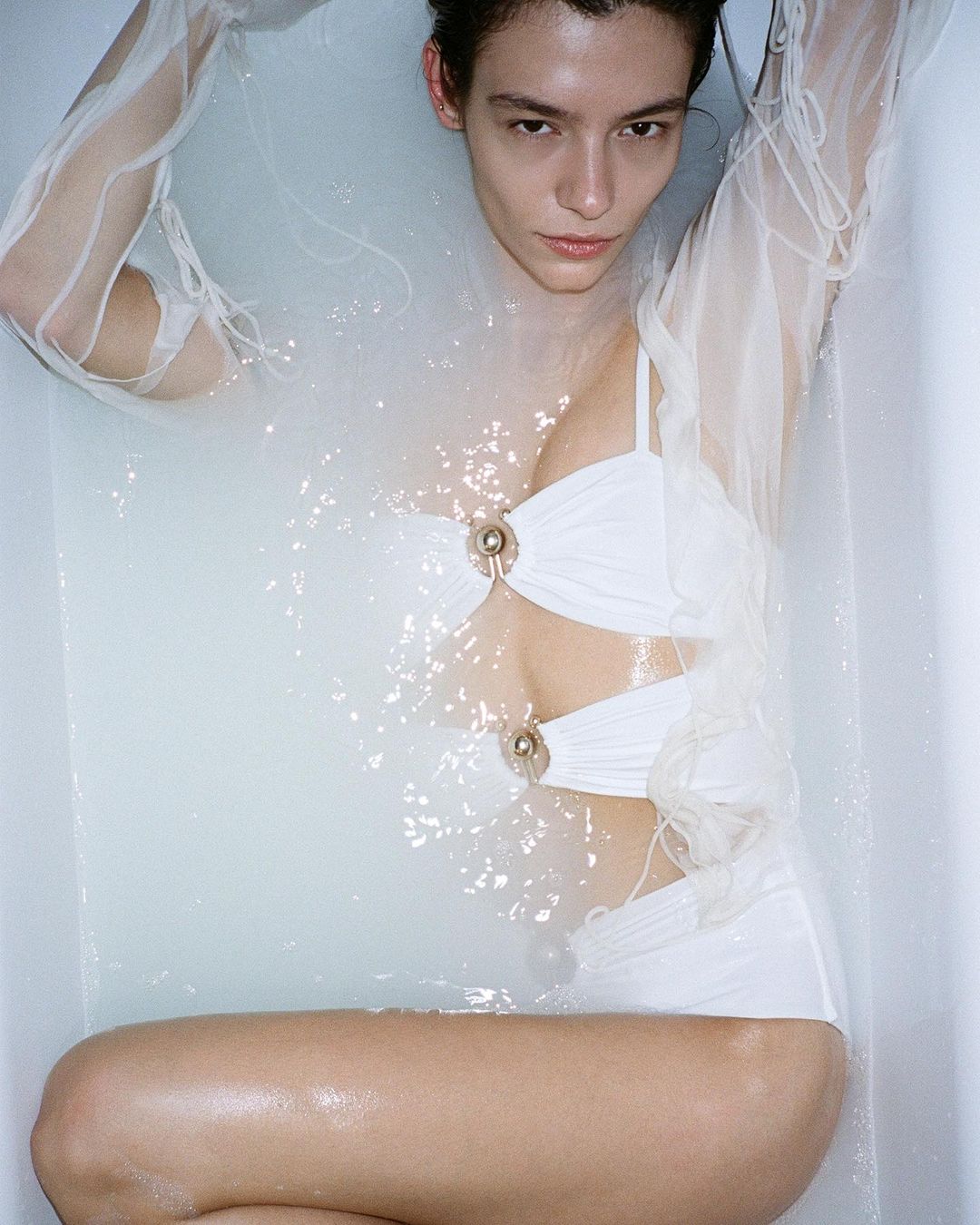 Brunette woman in white bathing suit poses in water-filled tub with arms at sides, wet hair, fair skin, minimalist look, and studio lighting.