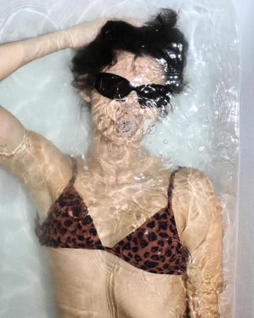 Woman in sunglasses and bikini top, submerged in water. Dark short hair. Relaxing in a bathtub, water bubbles. Clean and minimalist.