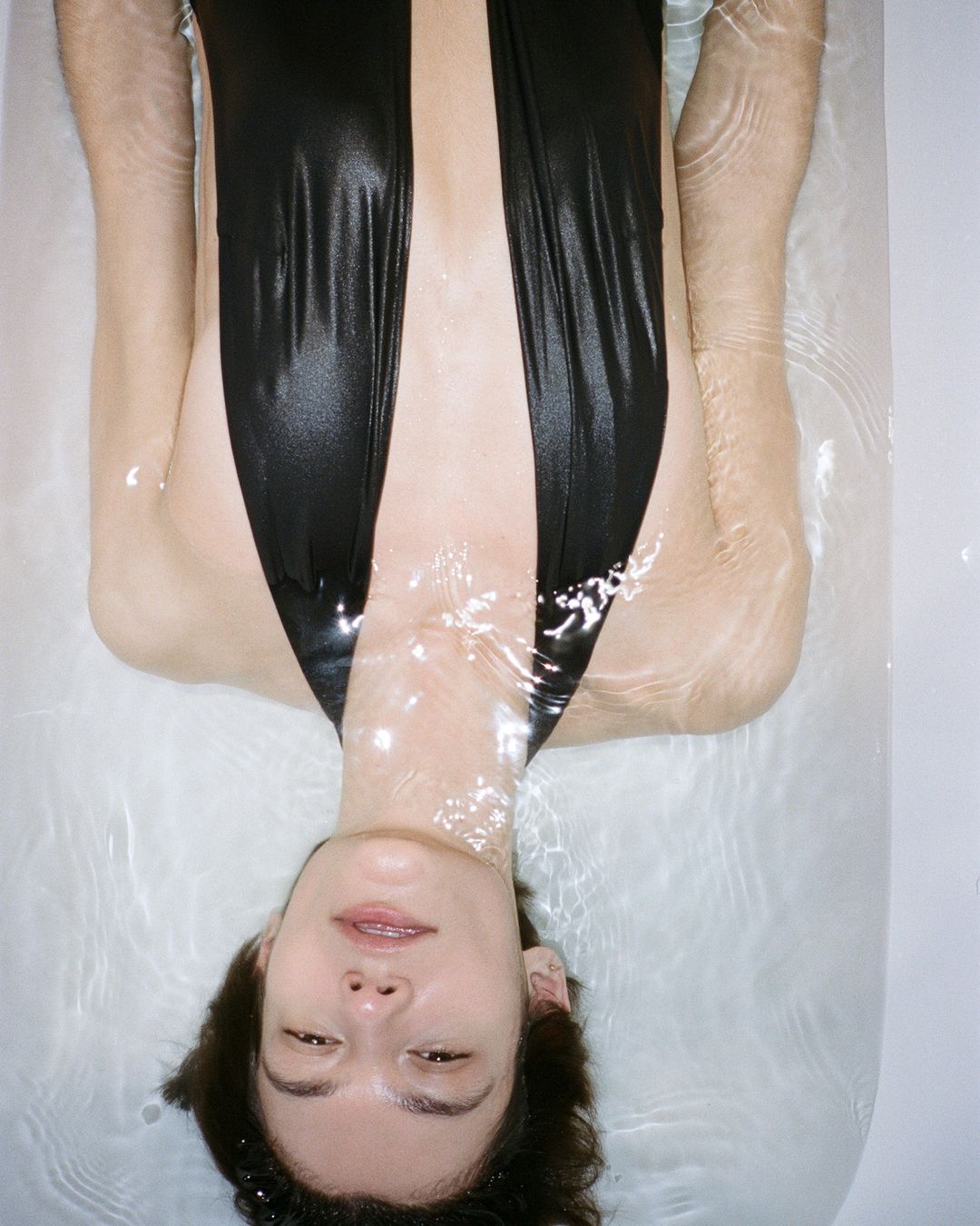 Model floating in a bathtub wearing a black plunging swimsuit for Christopher Esber Swim PF 24 campaign.