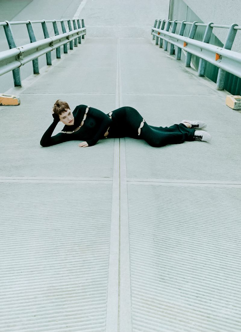 A woman lies on a concrete structure wearing a black outfit and light-colored sneakers, with metal barriers in the background.
