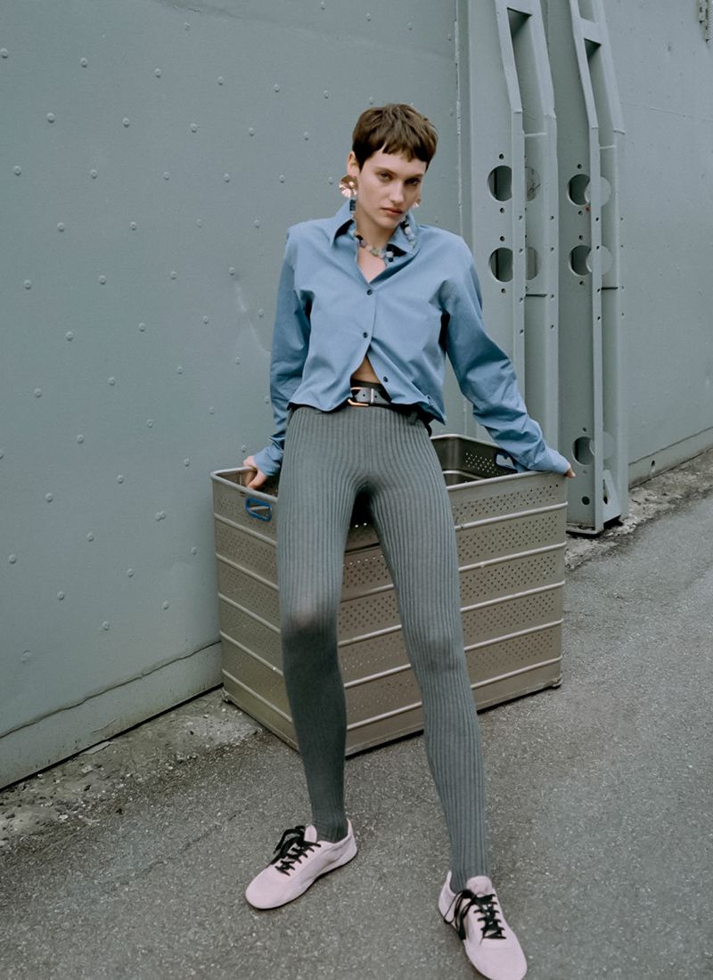Fashionable woman in a light blue shirt and ribbed leggings poses next to a metal box against a neutral grey background.