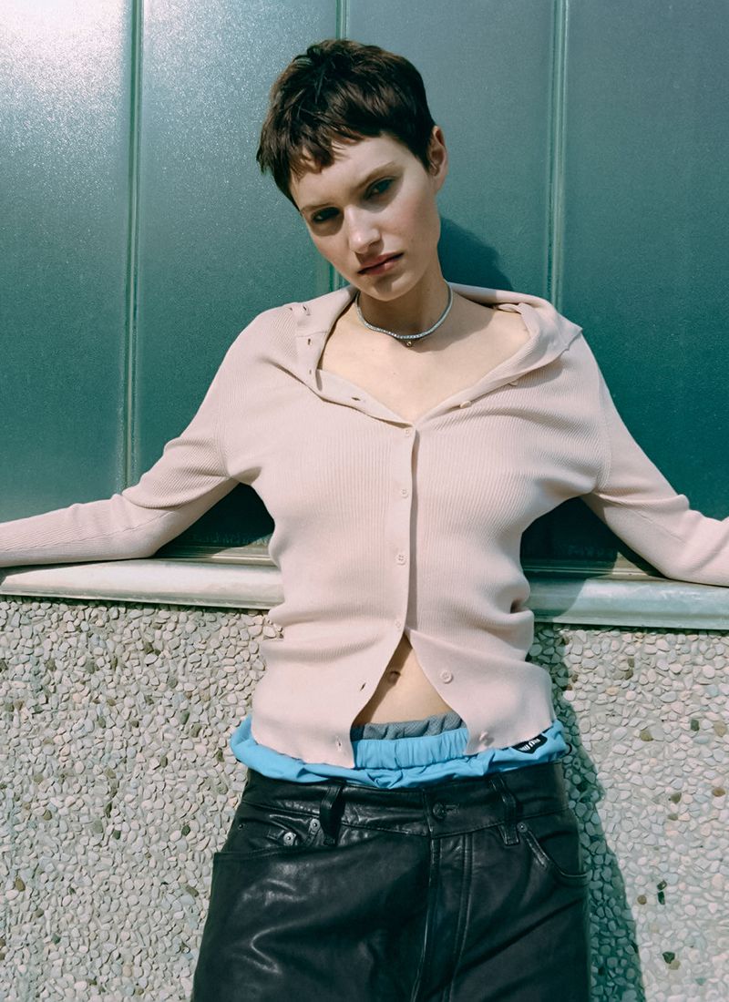 A young woman with short hair poses in front of a textured wall wearing a hooded cardigan and dark pants. Neutral outdoor lighting.