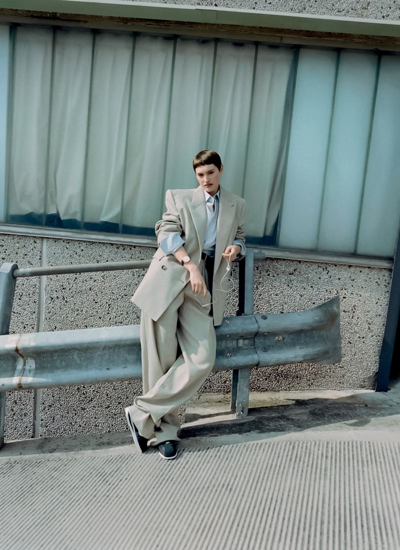 An adult standing in a beige suit leans against a metal barrier with a building and window in the background on a sunny day.