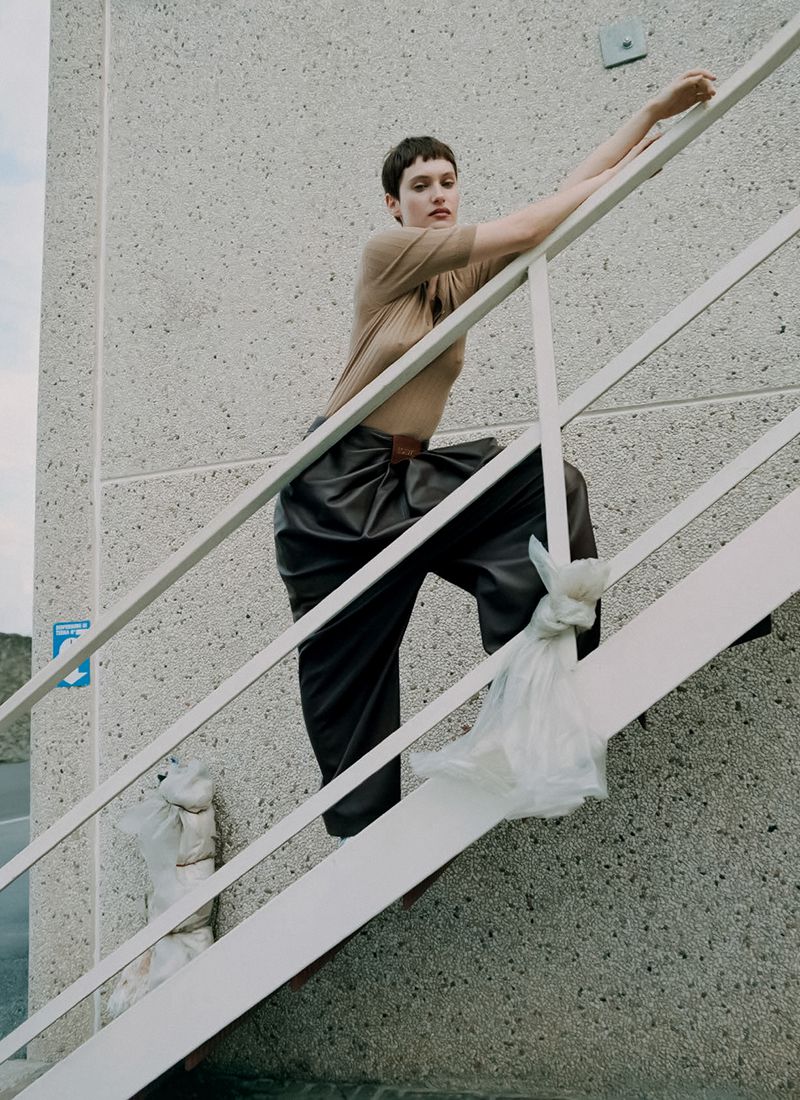 An adult female with short brunette hair poses on an outdoor staircase with plastic bags, wearing a tan top and dark wide-leg pants.