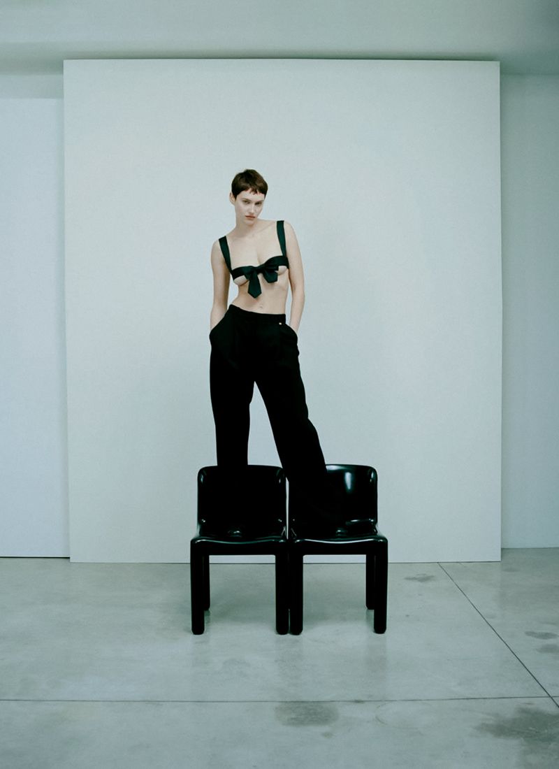 An adult female stands on two black chairs in a minimalist studio wearing a black bra top and trousers against a white backdrop.