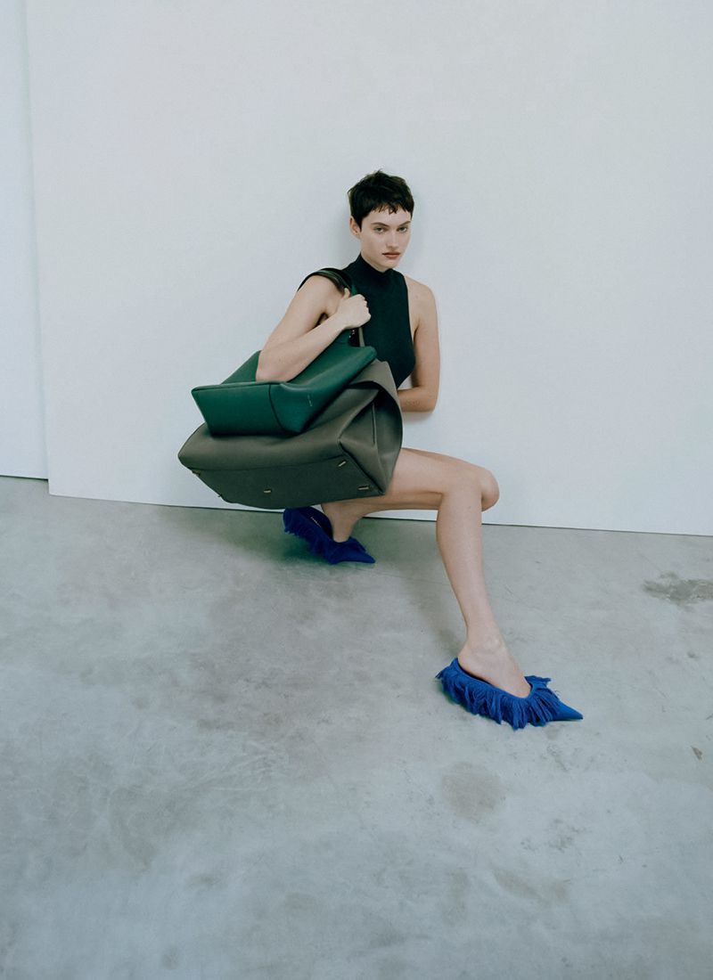 Fashion shot of a slender woman in a black top and blue shoes, kneeling, holding a green tote bag, against a white wall and grey floor.