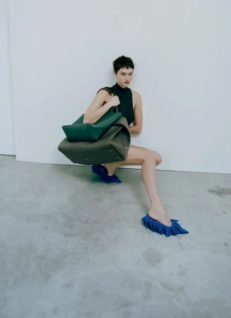 Fashion shot of a slender woman in a black top and blue shoes, kneeling, holding a green tote bag, against a white wall and grey floor.