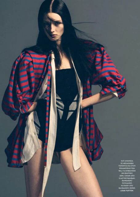 Fashion photo of a young adult woman with long black hair wearing a black bodysuit and a red and blue striped jacket in studio.