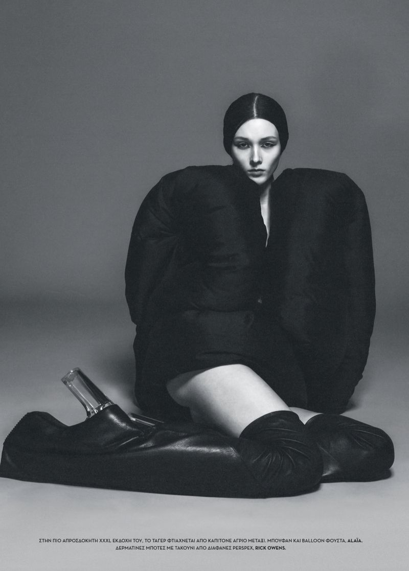 Fashion editorial shot of a woman in an oversized black jacket and heelless shoes against a gray backdrop in a studio setting.
