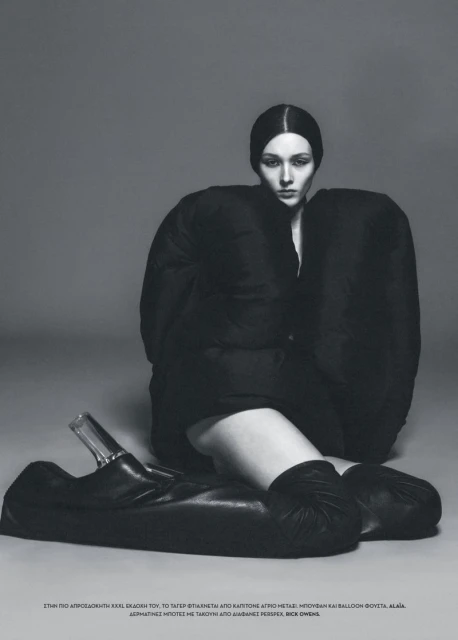 Fashion editorial shot of a woman in an oversized black jacket and heelless shoes against a gray backdrop in a studio setting.