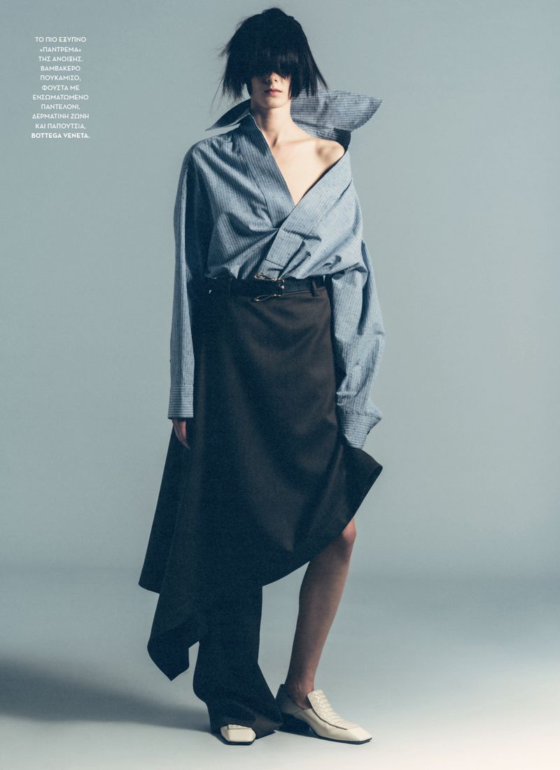 Model with dark fringed hair wearing an oversized blue striped shirt and dark skirt against a grey studio background