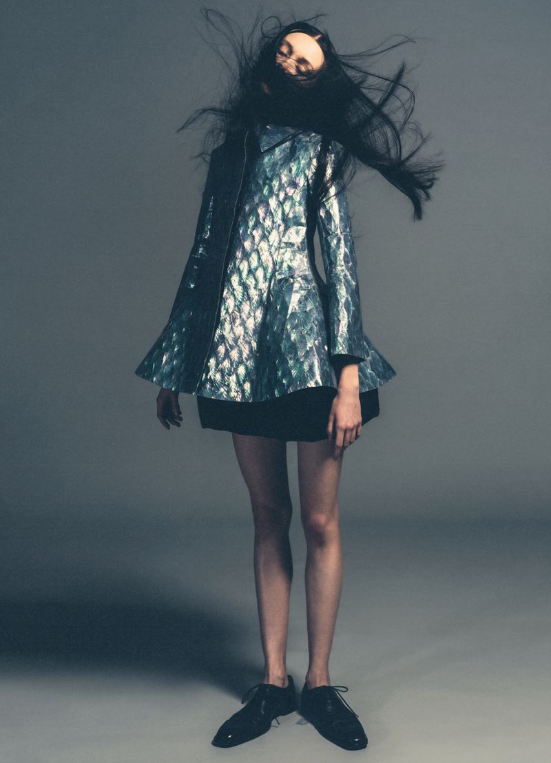 Fashion model in iridescent jacket with black skirt and patent-leather shoes poses on a gray studio background with flying hair.