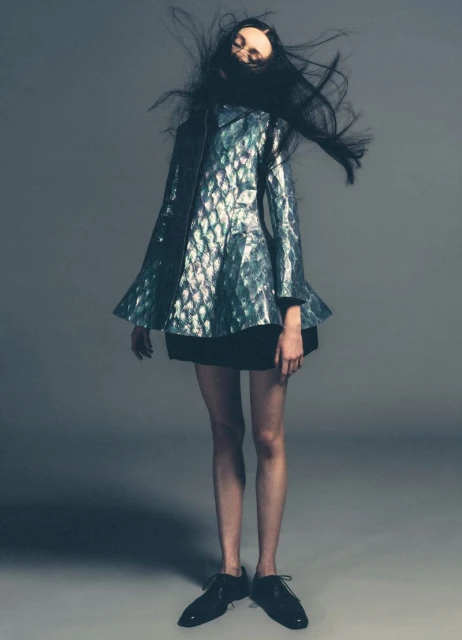 Fashion model in iridescent jacket with black skirt and patent-leather shoes poses on a gray studio background with flying hair.