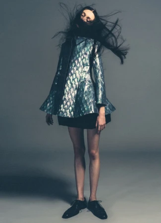 Fashion model in iridescent jacket with black skirt and patent-leather shoes poses on a gray studio background with flying hair.