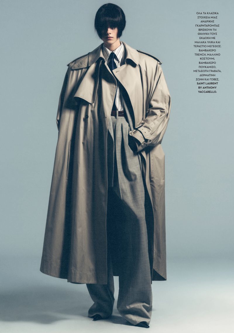 Model wearing an oversized beige trench coat over a striped shirt and grey trousers against a plain grey backdrop.