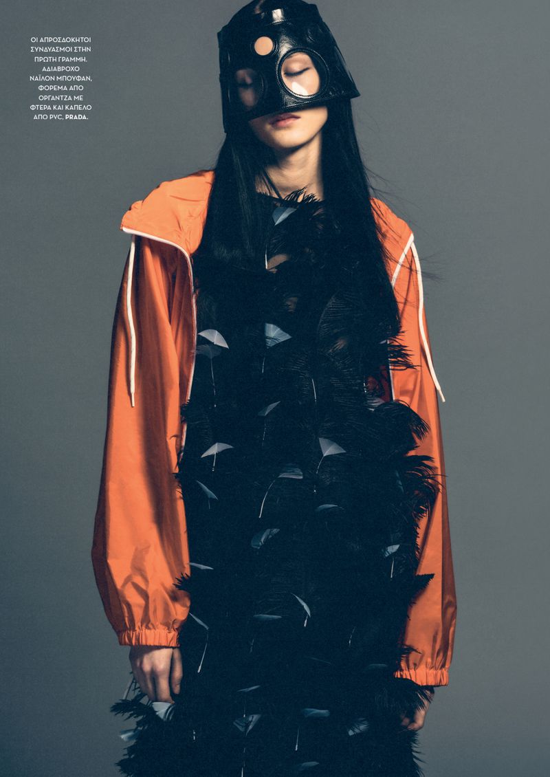 Fashion editorial shot of a model with long black hair, wearing a black feathered dress and orange jacket against a grey backdrop.