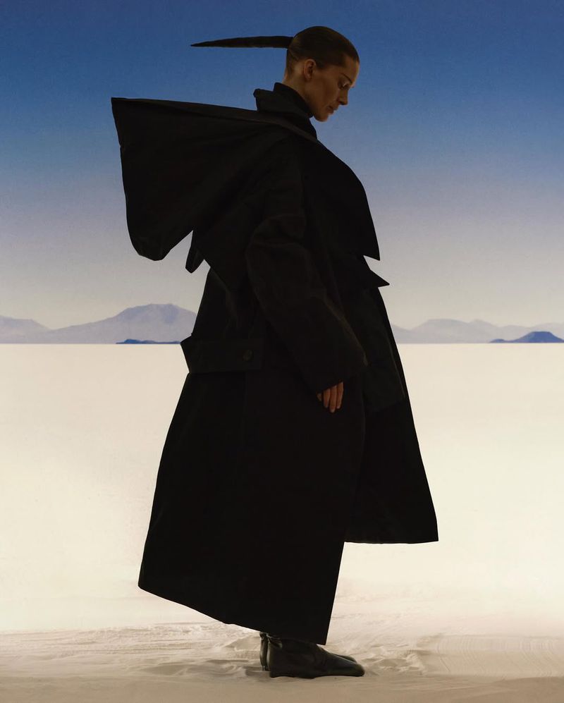 Side profile of a slender person in a black, oversized coat against a gradient blue sky with white mountains in the background.
