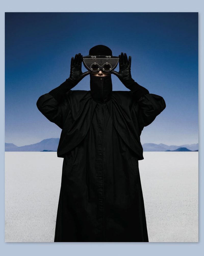 Person wearing black clothing and vintage goggles poses in a white, desert-like landscape under a blue sky. Fashion photography.
