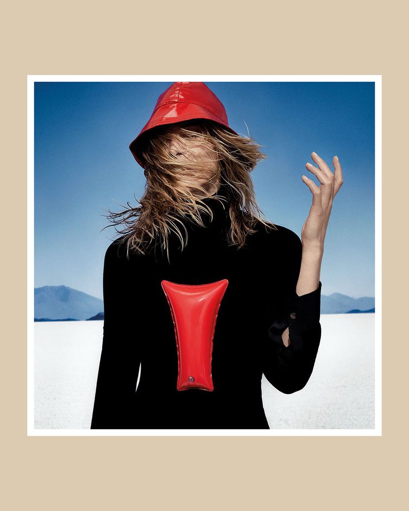 A person with blond hair stands outdoors on a salt flat wearing a black turtleneck and a red hat with a matching red inflatable.