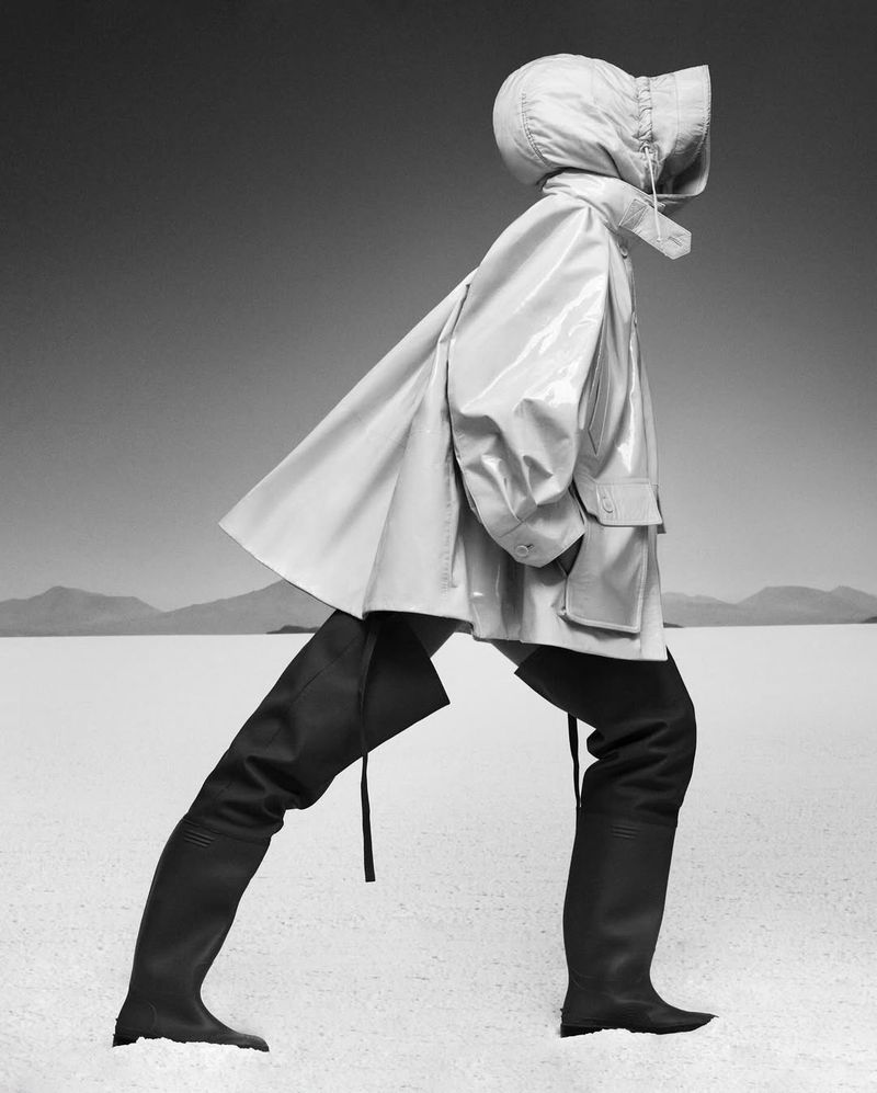 Person in hooded raincoat and knee-high boots strides across a bright white desert plain beneath a pale sky with distant mountains.