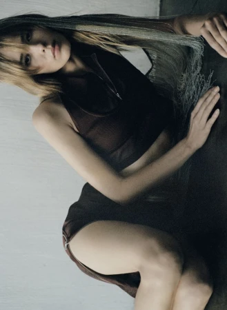 Woman with long blonde hair poses in a dark brown outfit with a zipper. She is laying against a white wall and a gray surface.