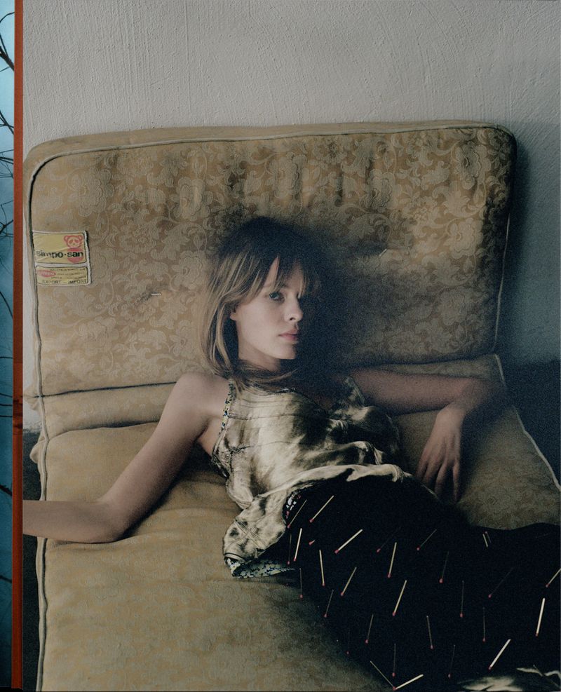 Adult woman with long blonde hair reclining on a vintage patterned cushion, wearing a patterned top and skirt with black background. Indoor.