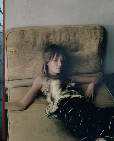Adult woman with long blonde hair reclining on a vintage patterned cushion, wearing a patterned top and skirt with black background. Indoor.