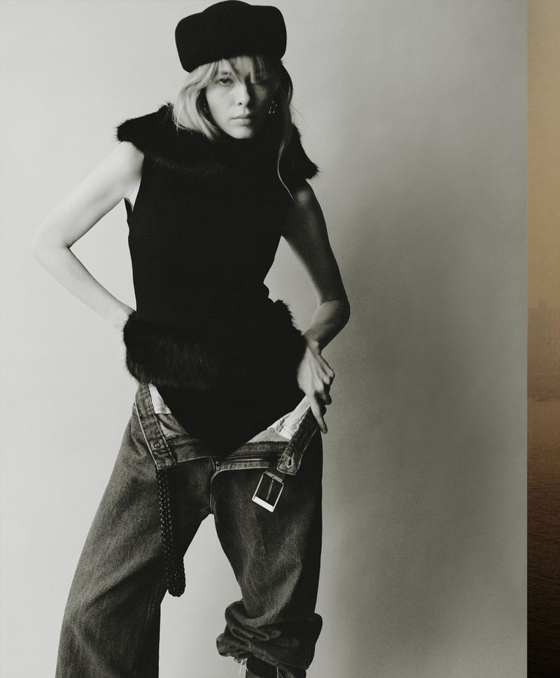 Monochrome fashion portrait of a woman with blonde hair wearing a black top, hat, and blue jeans, posing against a neutral background.