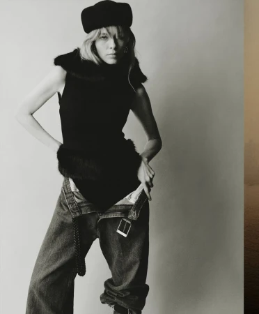 Monochrome fashion portrait of a woman with blonde hair wearing a black top, hat, and blue jeans, posing against a neutral background.