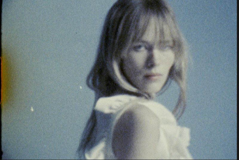 Blonde woman in white top looks over her shoulder against a soft blue background with vintage film aesthetic and shallow depth of field.