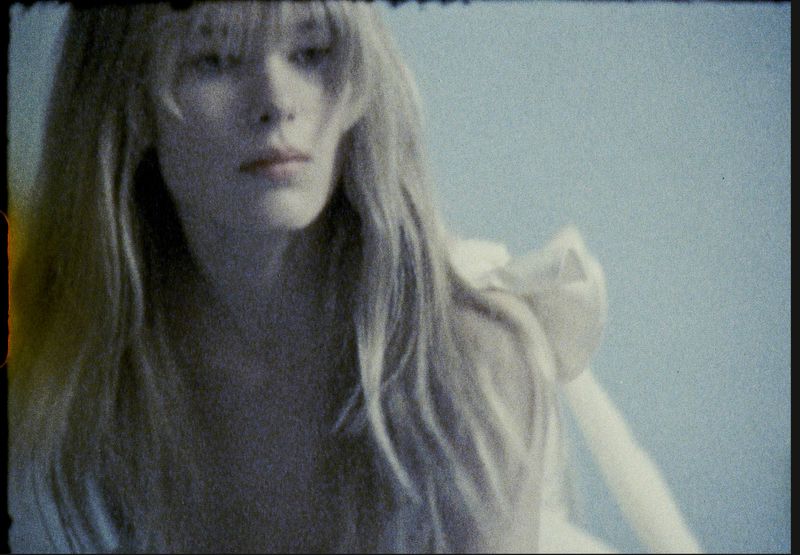 Adult female with blonde hair gazes to the side in a medium shot against a plain light-blue background in a portrait-oriented frame.