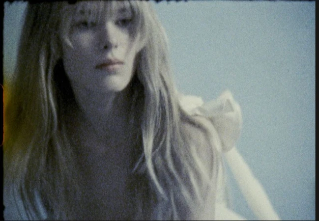Adult female with blonde hair gazes to the side in a medium shot against a plain light-blue background in a portrait-oriented frame.