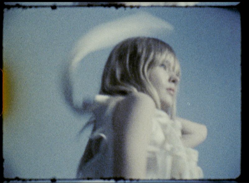 Portrait of a young woman with blonde hair, wearing a white ruffled top, posing against a blue sky. Soft focus with retro aesthetic.