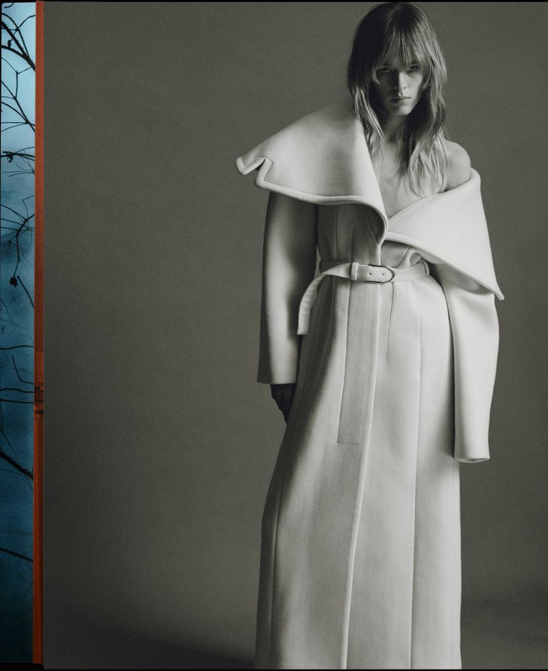 A woman stands in a photo studio wearing an off-white, belted high-fashion coat with an exaggerated collar against a grey backdrop.