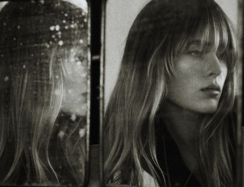 Monochrome portrait of a woman with bangs, reflected in a mirror, capturing an intimate and reflective moment in desaturated tones.