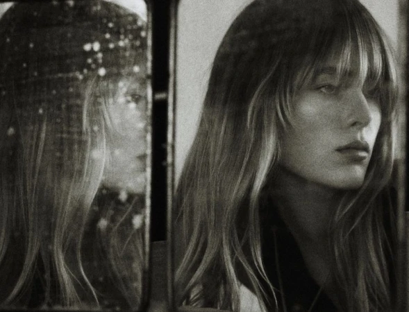 Monochrome portrait of a woman with bangs, reflected in a mirror, capturing an intimate and reflective moment in desaturated tones.