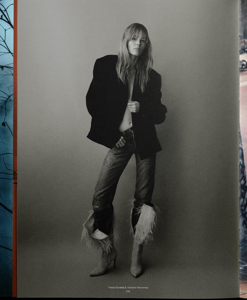 Monochrome fashion portrait of woman in oversized blazer, sparkly jeans with feather trim, and heels posing in a studio setting.