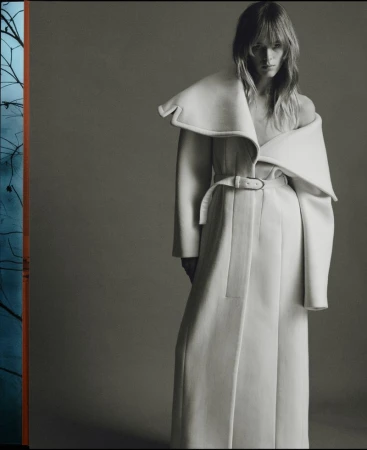 Model Kristine Lindseth wearing a long white belted coat with an asymmetric collar against a plain grey studio backdrop.